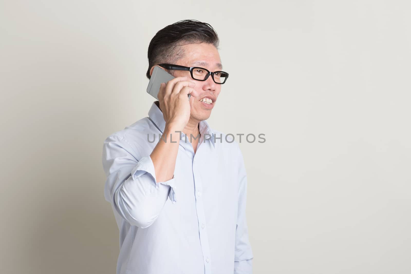 Portrait of single mature 50s Asian man in casual business talking on smartphone and smiling, standing over plain background with shadow. Chinese senior male people.