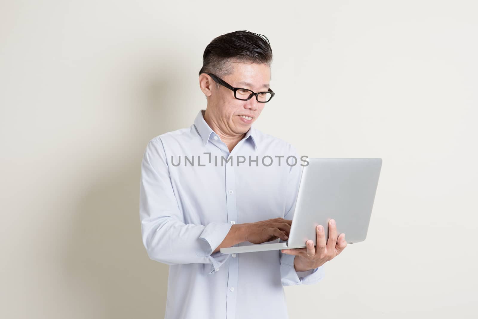 Portrait of single mature 50s Asian man in casual business using laptop computer and smiling, standing over plain background with shadow. Chinese senior male people.