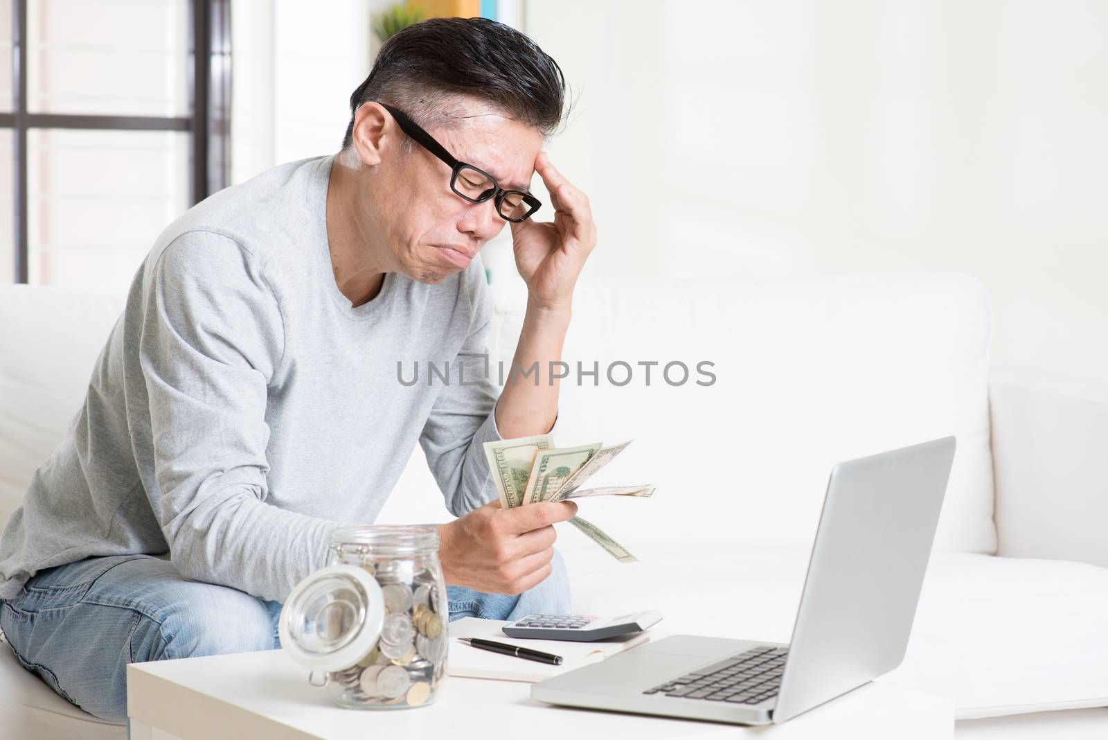 Financial problem concept. Portrait of 50s mature Asian man counting money with worried expression, sitting on sofa at home.