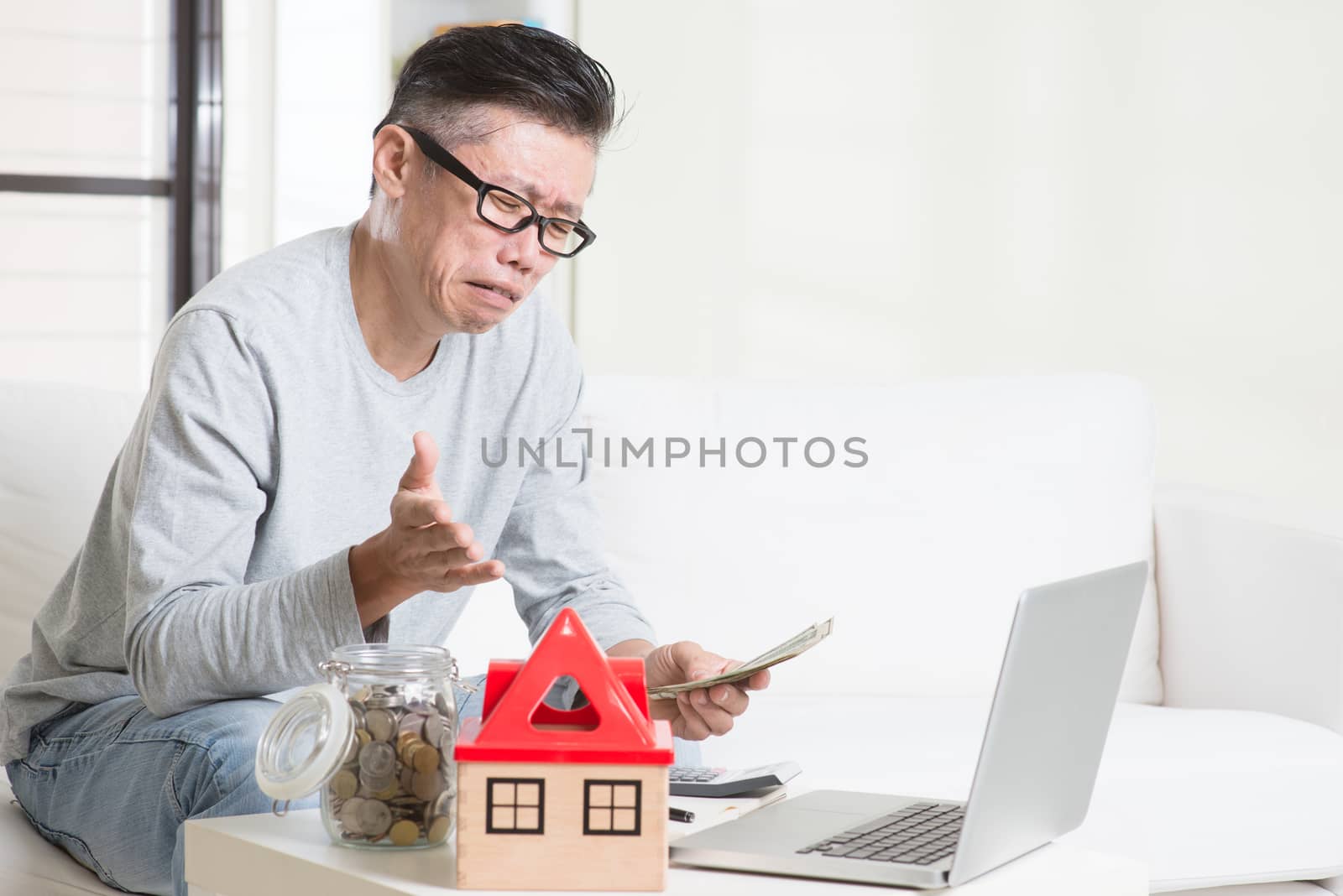 Cash flow problem concept. Portrait of 50s mature Asian man counting money with worried expression, sitting on sofa at home.