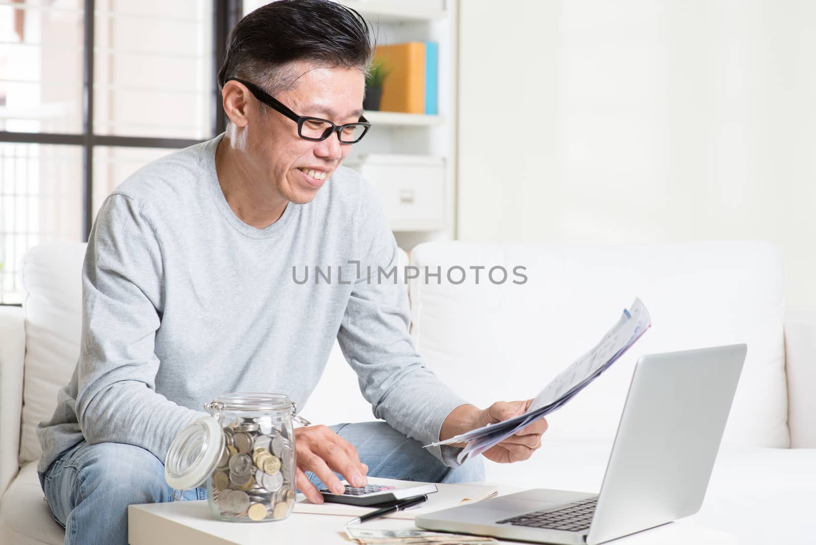 Portrait of 50s mature Asian man looking at bill and laptop in the living room. Saving, retirement, retirees financial planning concept.