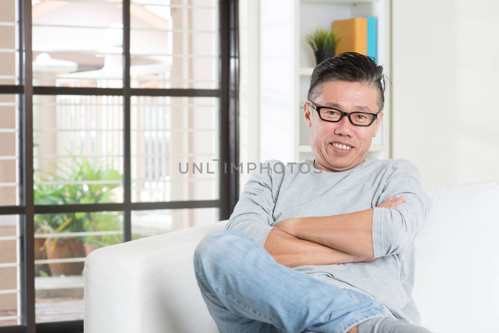 Portrait of happy mature 50s Asian man smiling and sitting at home. Senior Chinese male relaxed and seated on sofa indoor.