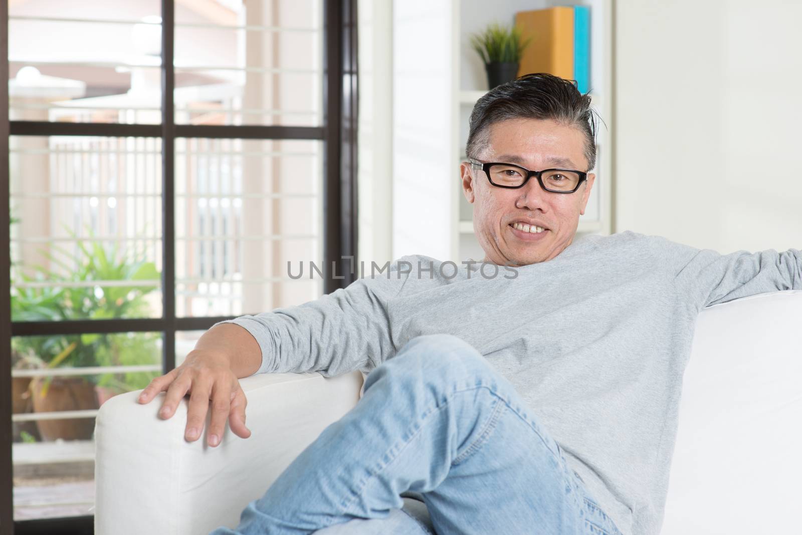 Portrait of mature 50s Asian man smiling and sitting at home. Senior Chinese male relaxed and seated on sofa indoor.