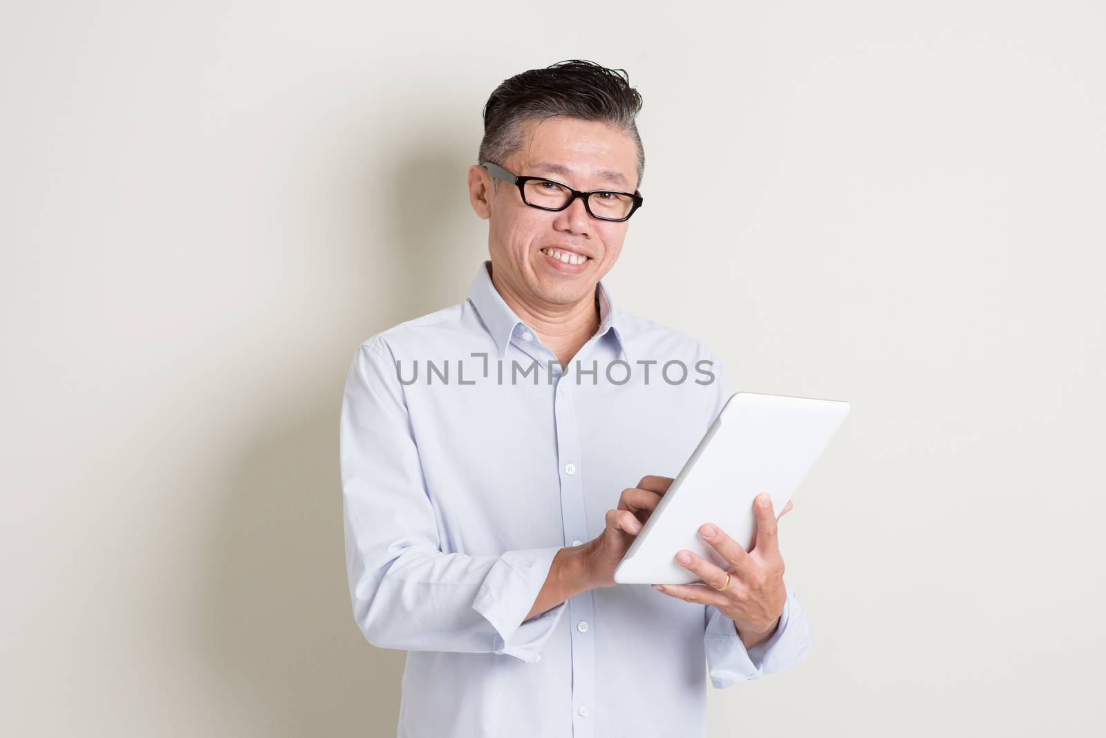 Portrait of single mature 50s Asian man in casual business using digital tablet pc and smiling, standing over plain background with shadow. Chinese senior male people.