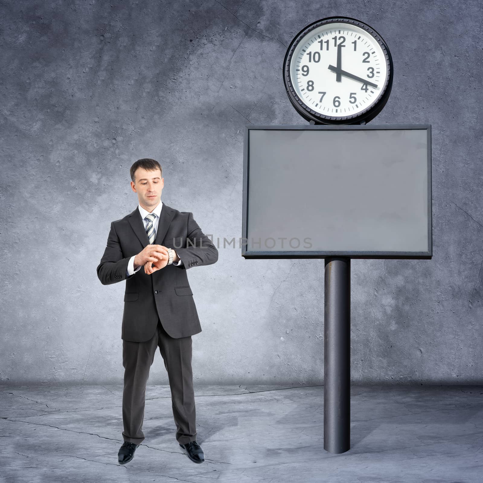 Businessman with looking at his watch near big clock on grey wall background