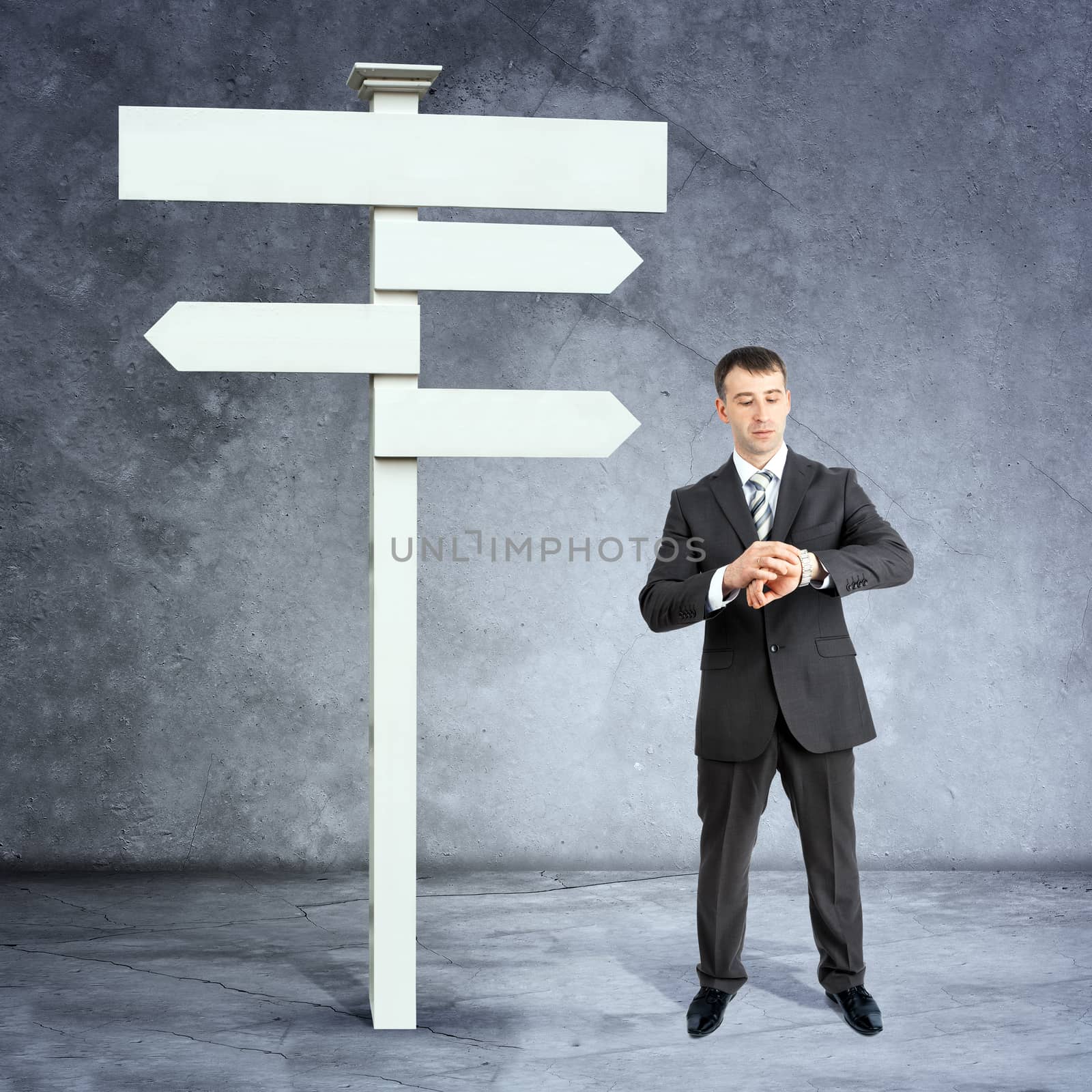 Businessman with looking at his watch near road sign on grey wall background