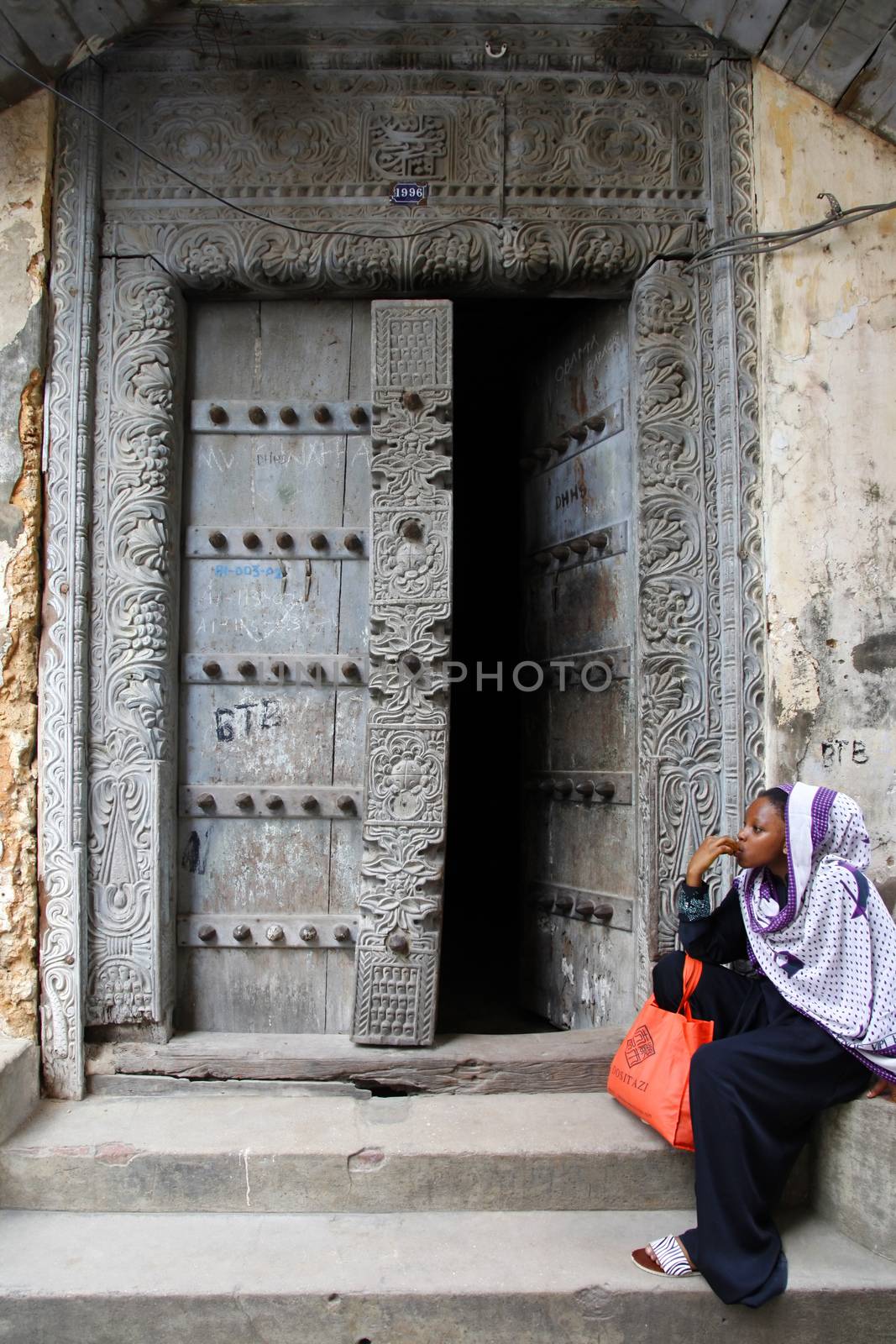 Stone Town, Tanzania - January 1, 2016: Traditional house with old door and woman in traditional Muslim clothes sitting on the street of Stone Town.