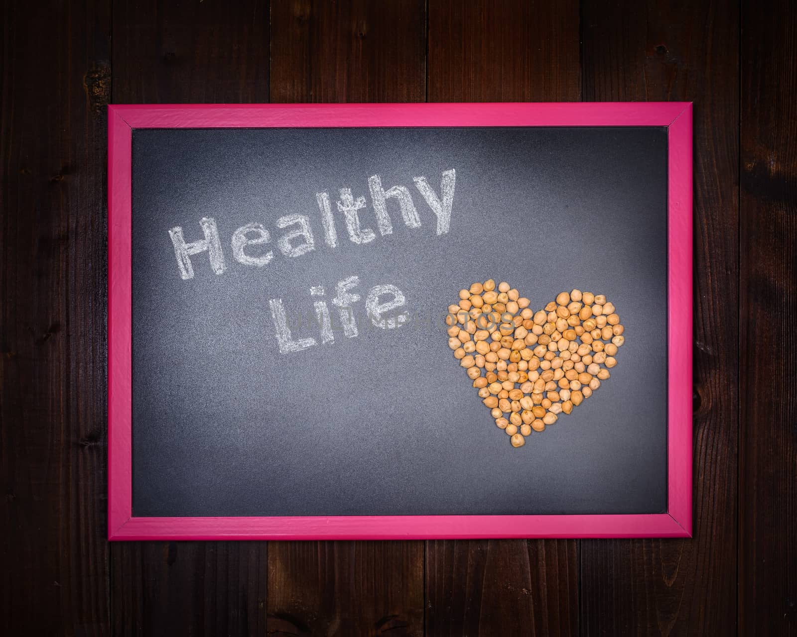 In the picture a blackboard, on the left side with written "Healthy Life" and chickpeas on the right side, drawing a heart on wooden background