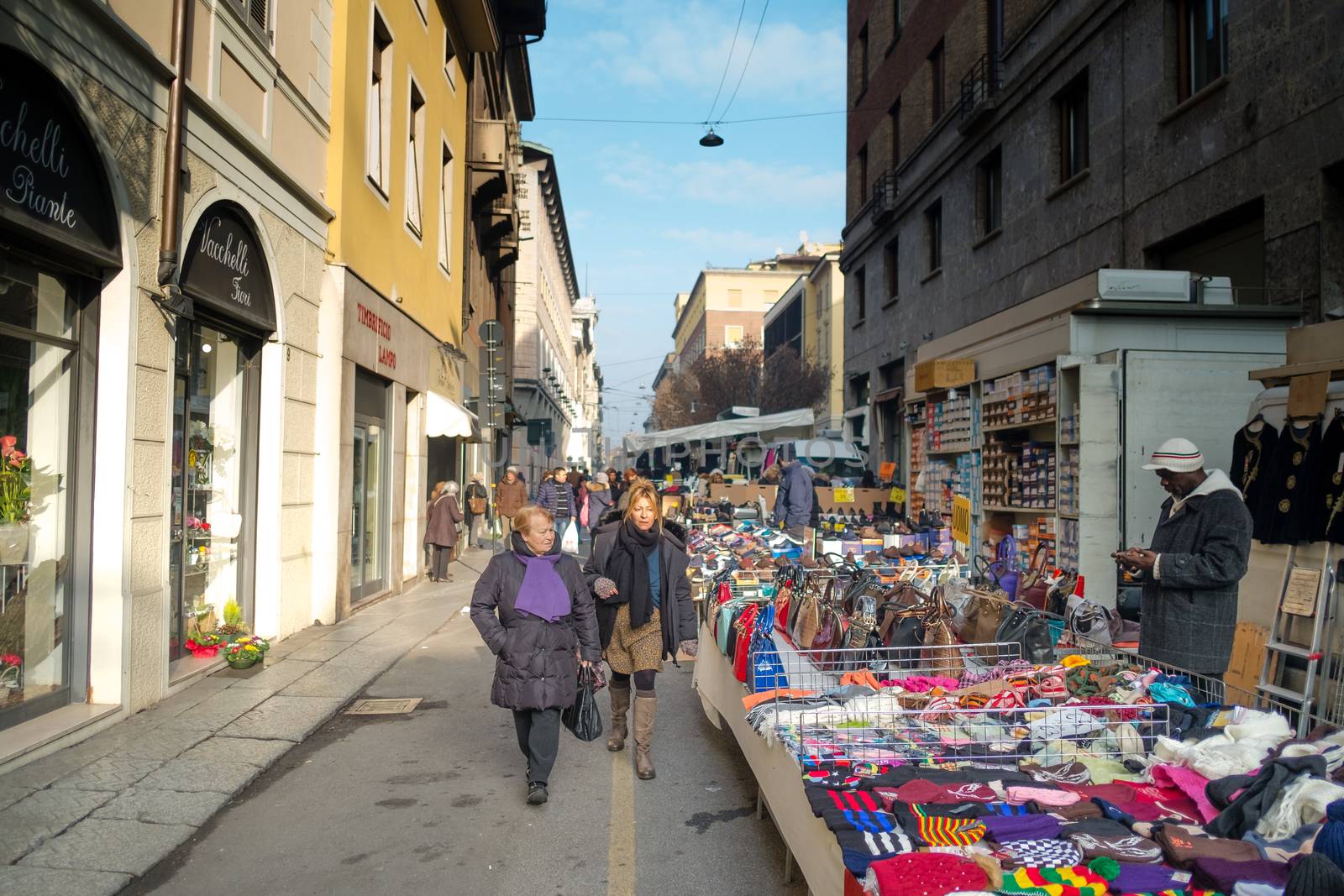 two women walking through the street market, and a vendor . Cremona market , january 2016