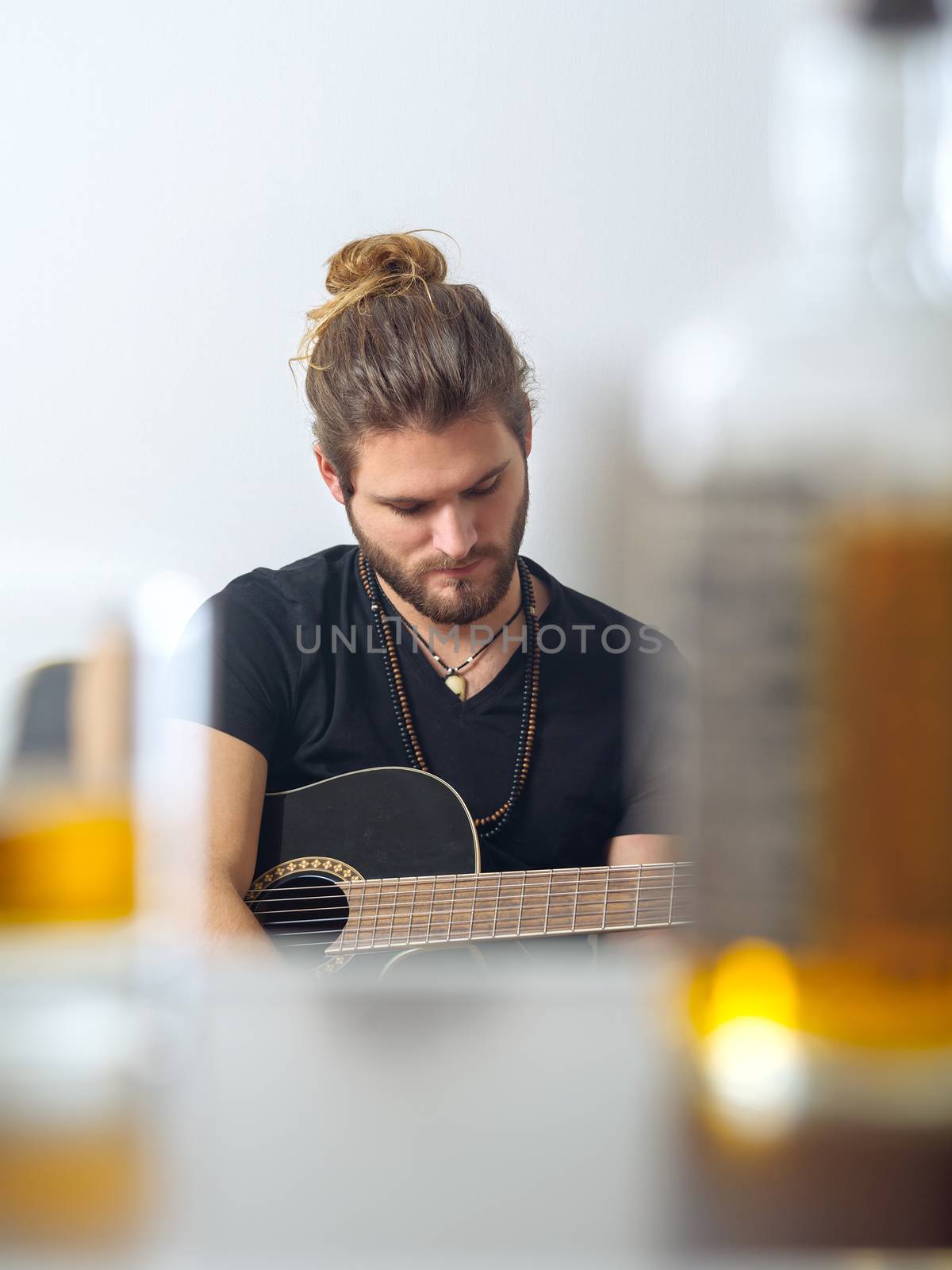 Photo of a male songwriter with acoustic guitar taken between a blurred whisky bottle and rocks glass.