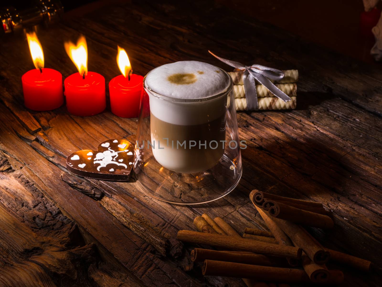 Frothy, layered cappuccino in a clear glass mug. Three red candles, cinnamon sticks and sweets. Romantic concept