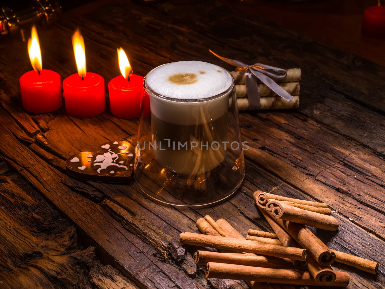 Frothy, layered cappuccino in a clear glass mug. Three red candles, cinnamon sticks and sweets. Romantic concept