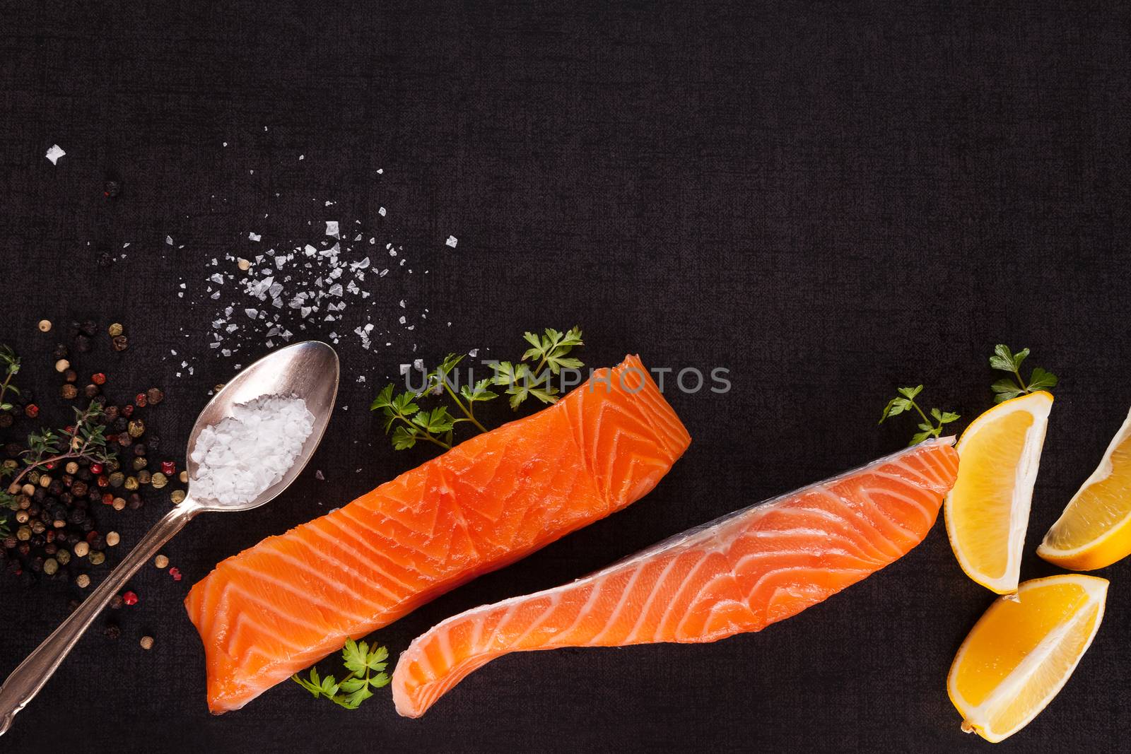 Salmon background with copyspace. Salmon steak, lemon, rosemary and parsley leaf and salt on black background, flat lay.
