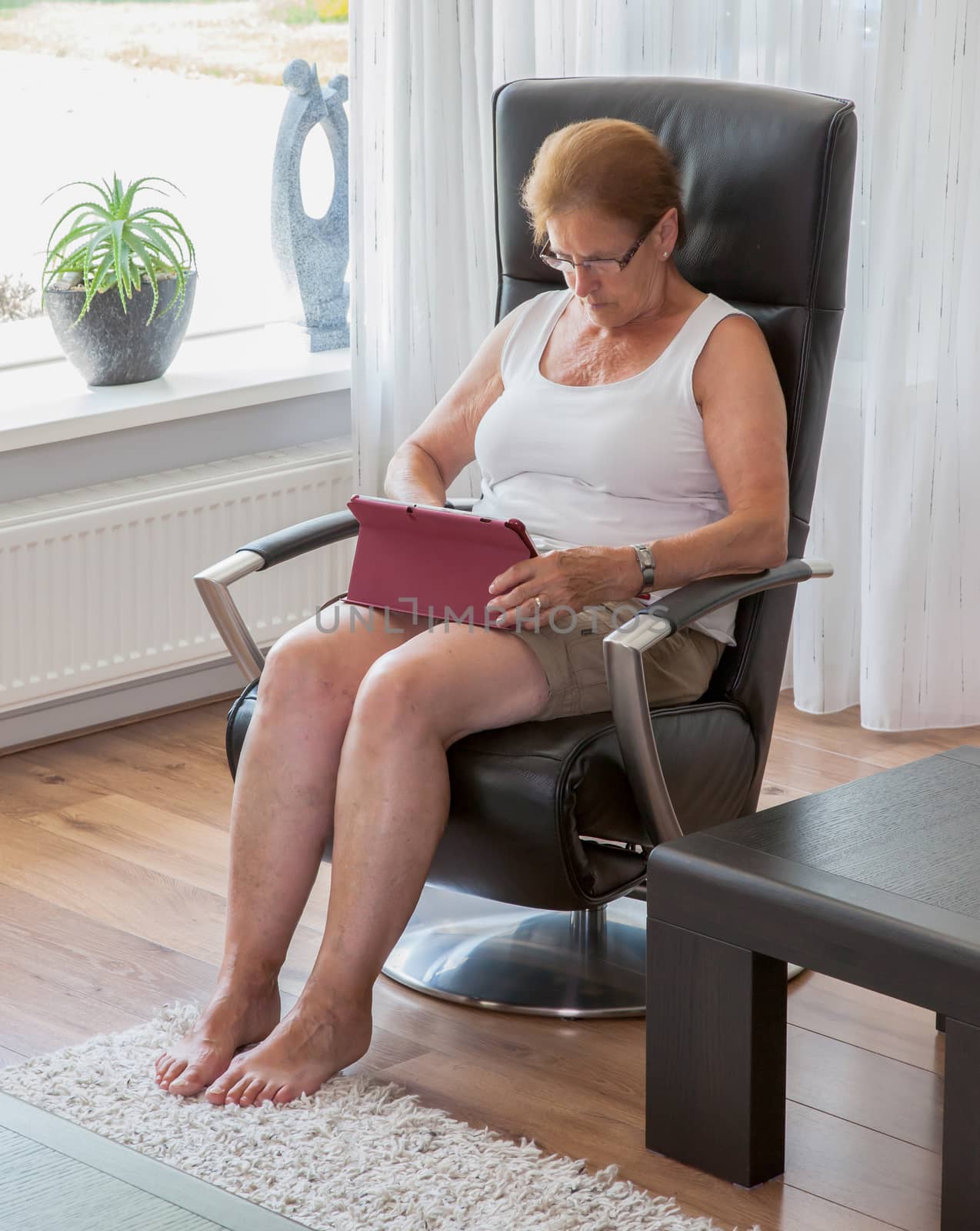 Senior woman sitting in a comfortable armchair in her living room concentrating on surfing the internet on a tablet