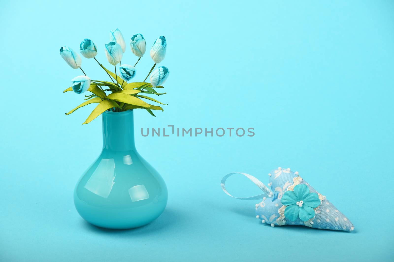 One small textile toy heart and mulberry silk tulip flowers bouquet in glass vase on tender blue design paper background