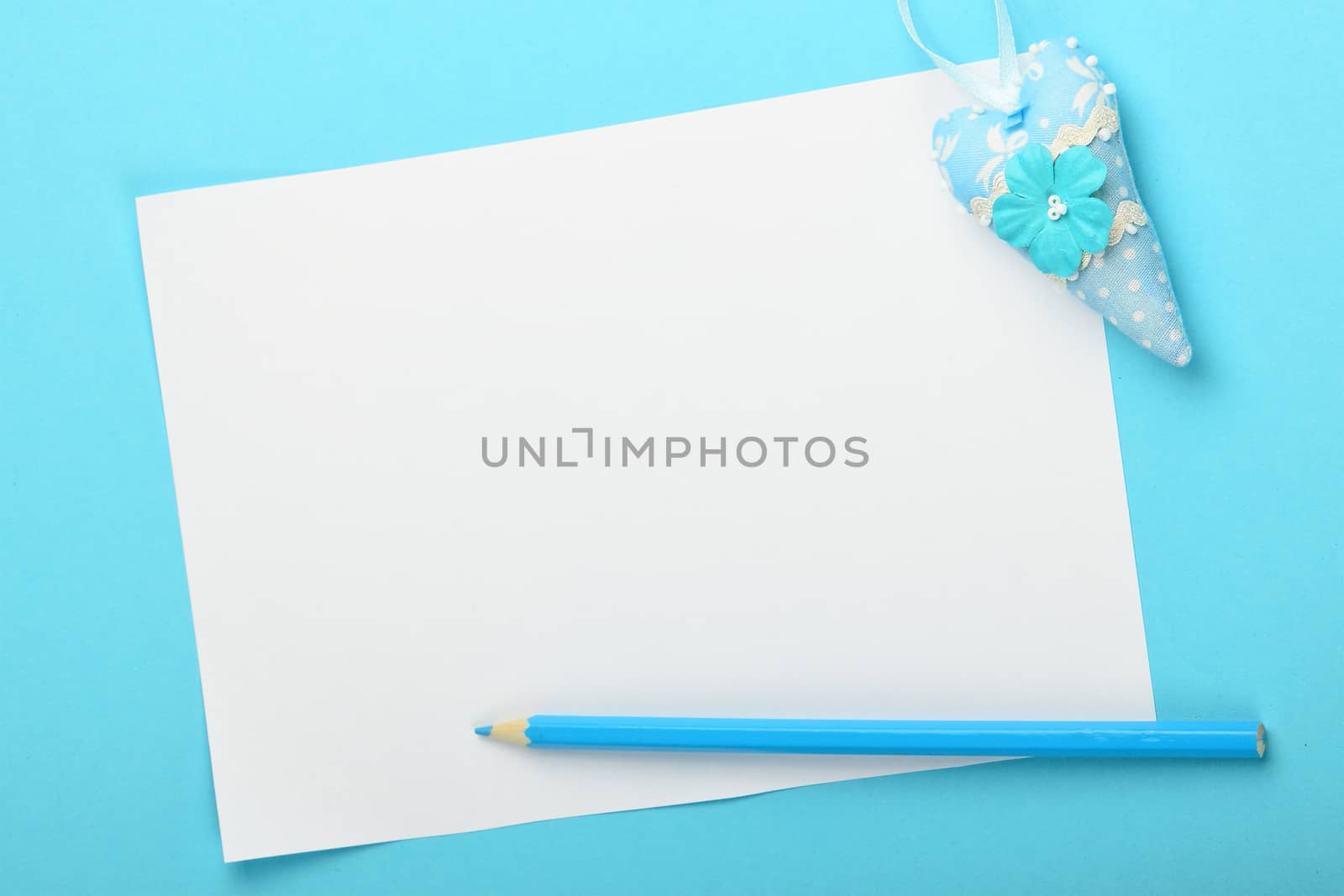 White paper note with small textile toy heart and pencil on tender blue background