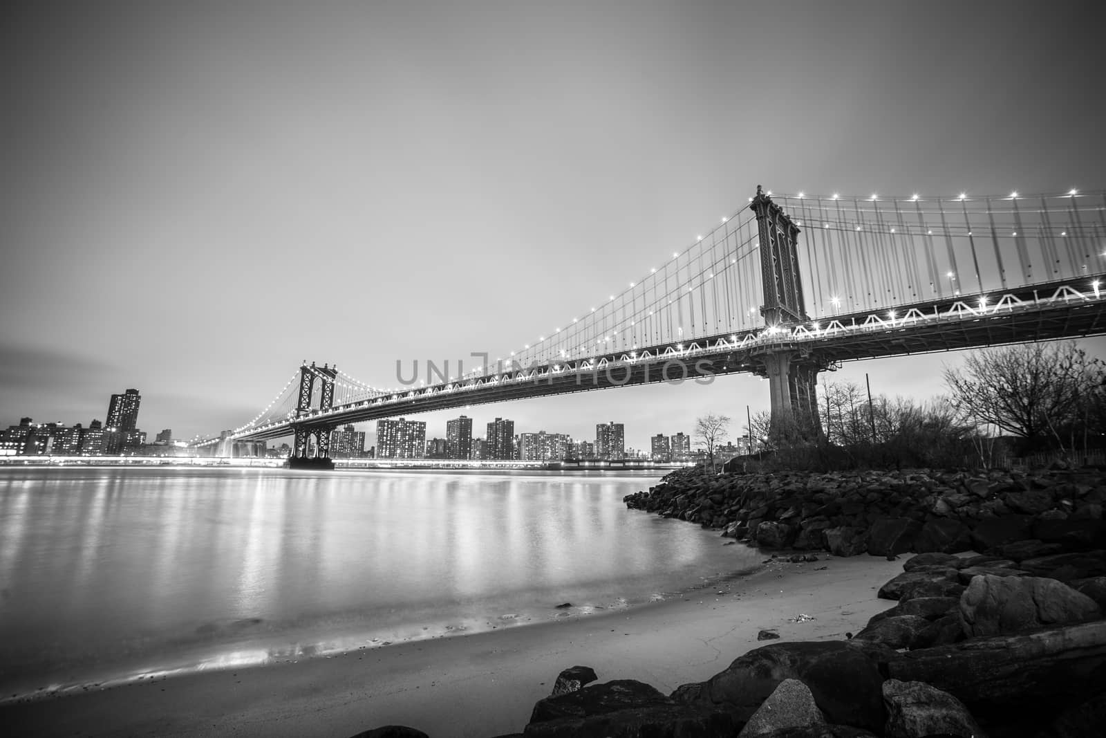 Manhattan bridge and New York City downtown skyline at dusk with skyscrapers illuminated over East River panorama. Black and white image.