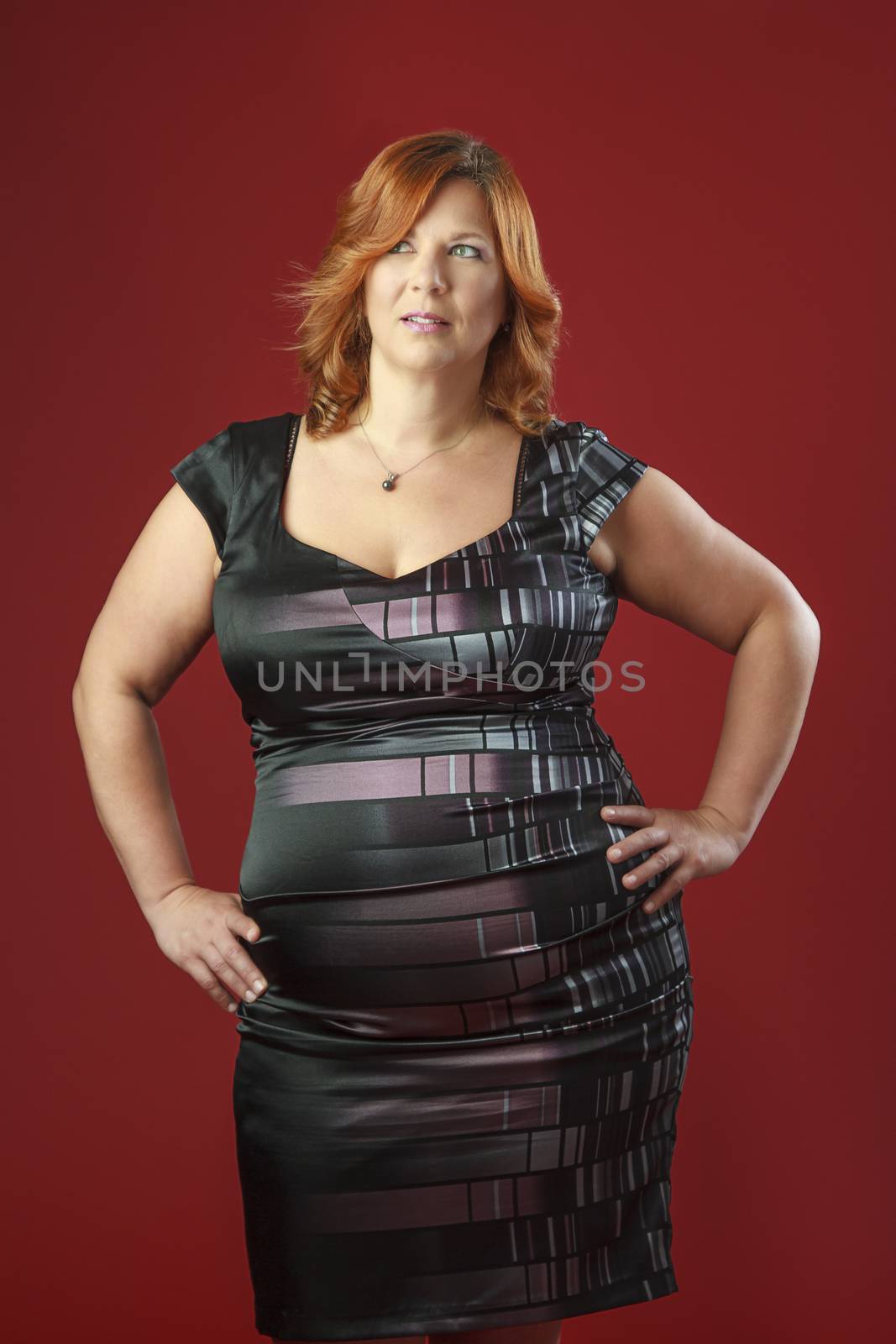 red hair woman, with her hand on her hips, wearing a cocktail dress against a red background