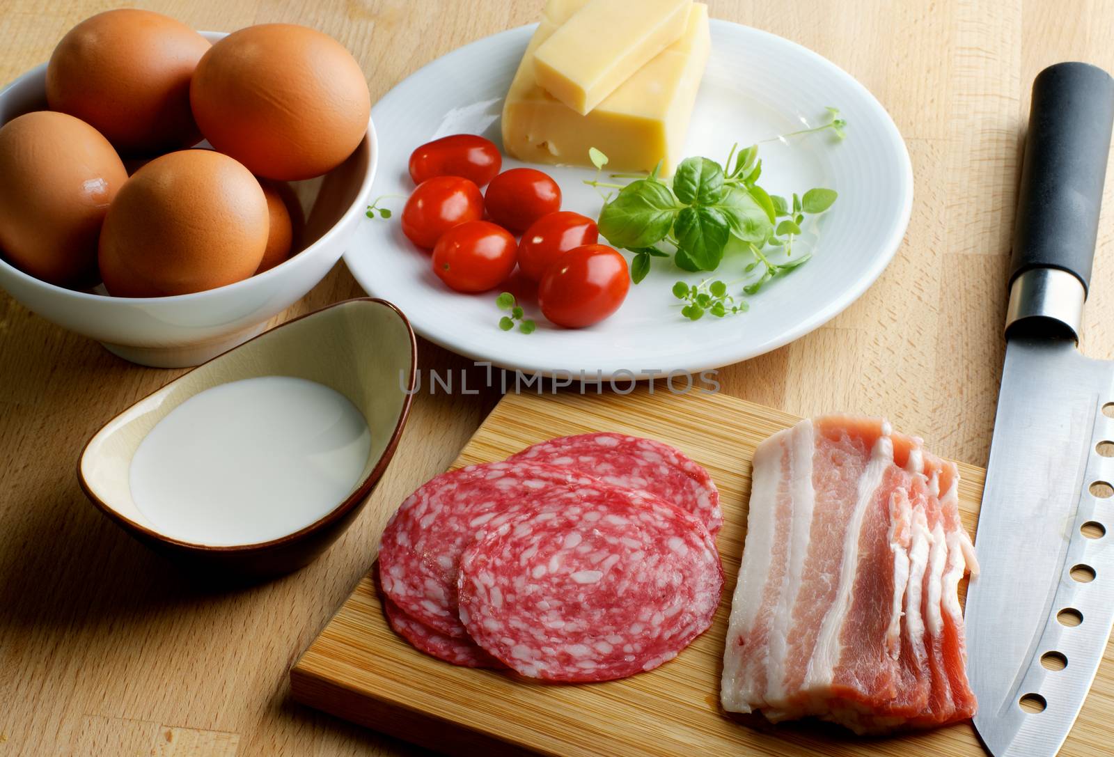 Ingredients of Omelet with Eggs, Milk, Cherry Tomatoes, Sausage, Bacon and Greens in Bowls with Kitchen Knife on Wooden Cutting Board closeup