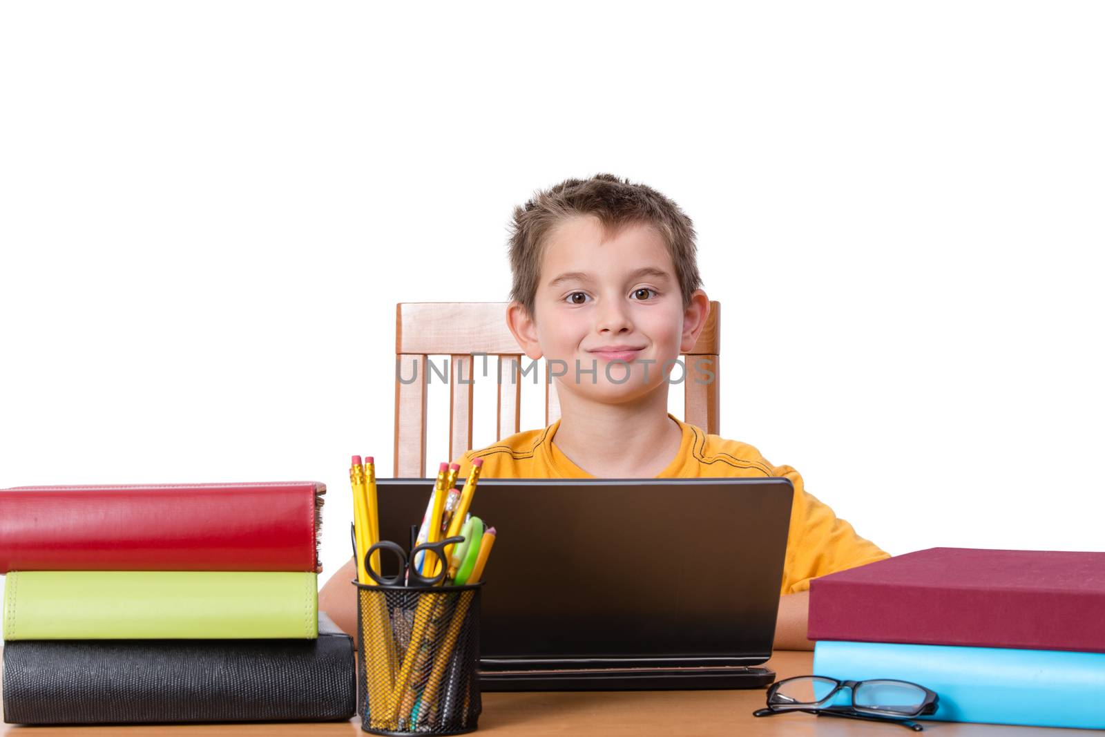 Smiling little boy at desk with laptop in between piles of large books and cup of pencils and pens