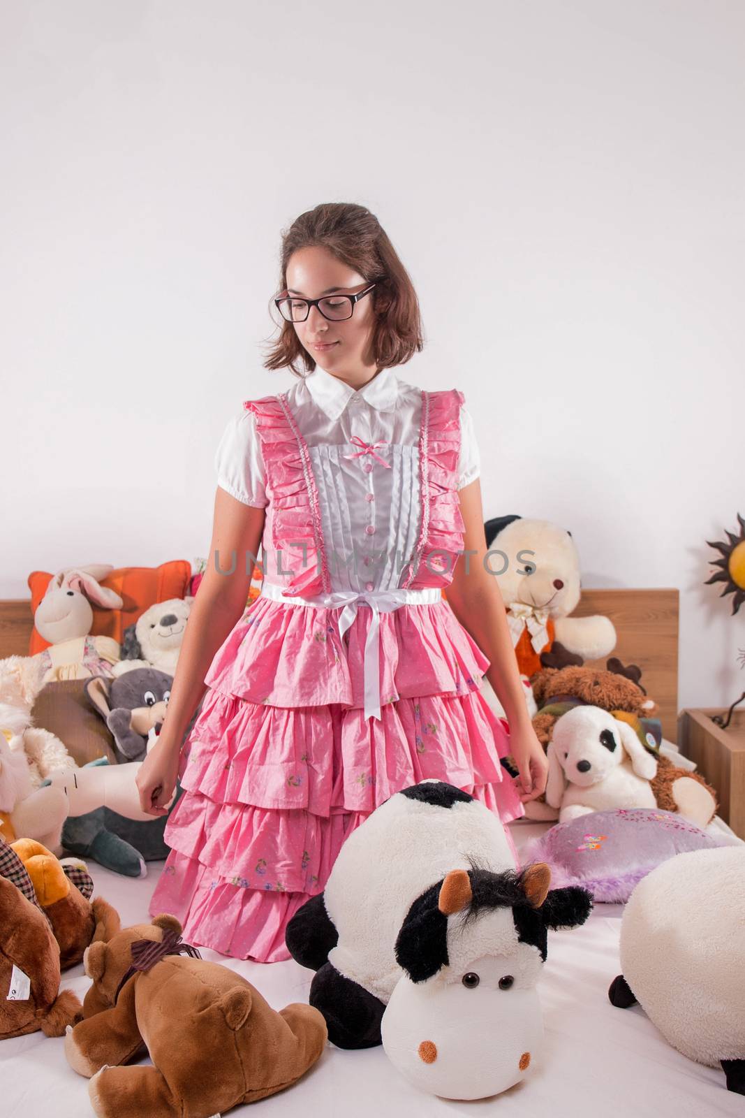 View of a young girl in a bedroom in a pink cute dress surrounded by stuffed toys.