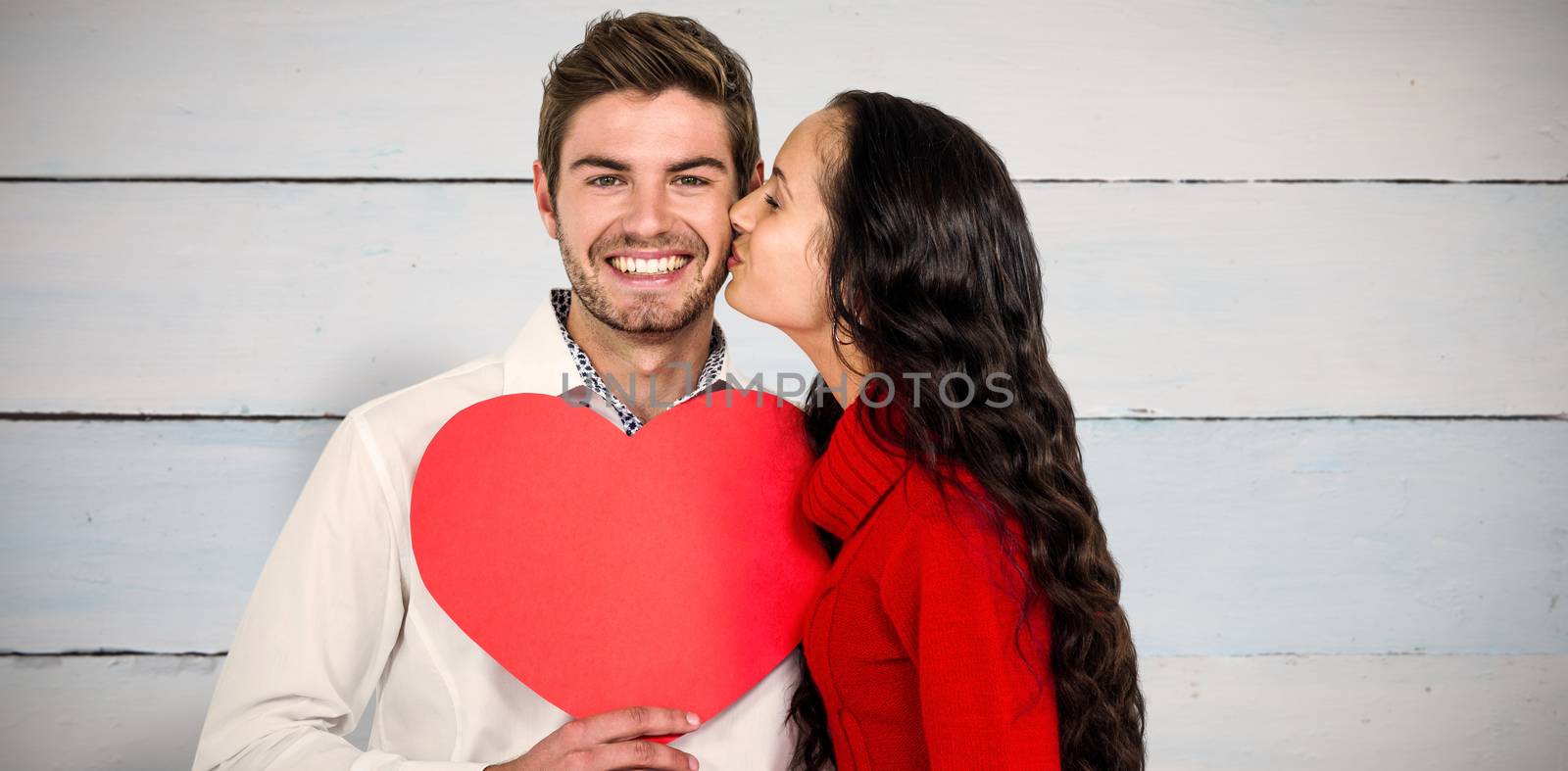 Man holding paper heart and being kissed by girlfriend against painted blue wooden planks