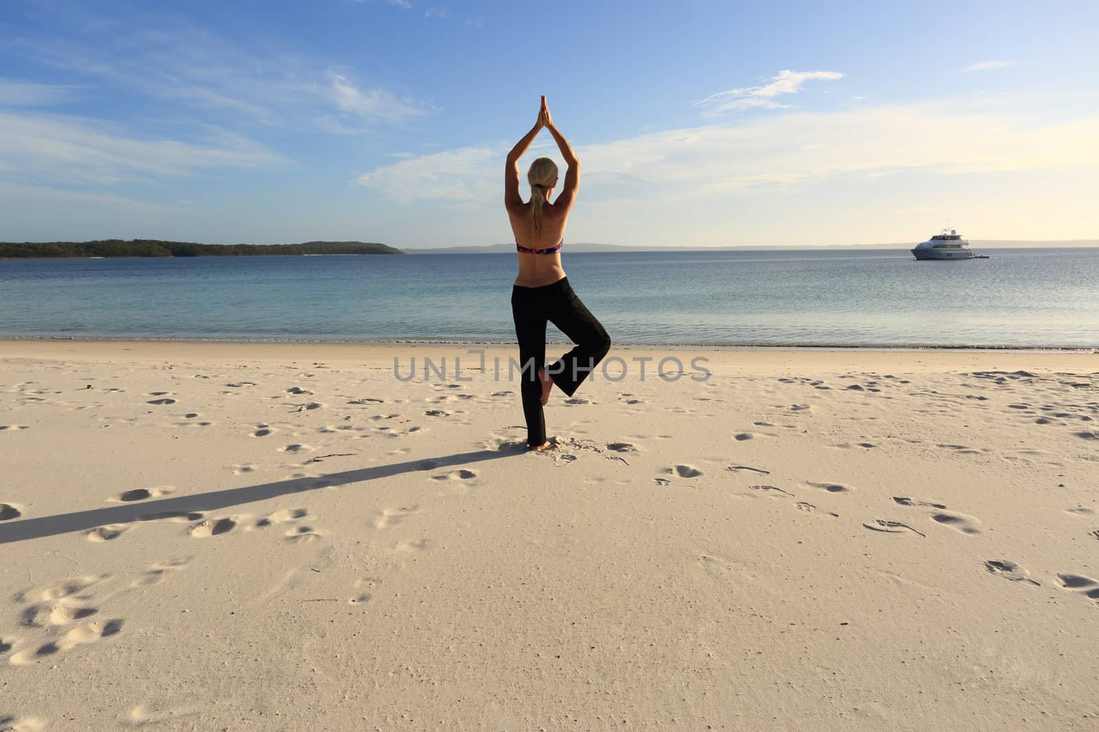Woman balanced in a yoga pose standing barefoot on one leg with her hands joined above her head at the beach in late afternoon sunlight, drawing long shadows on the sand. Asana vrksasana.