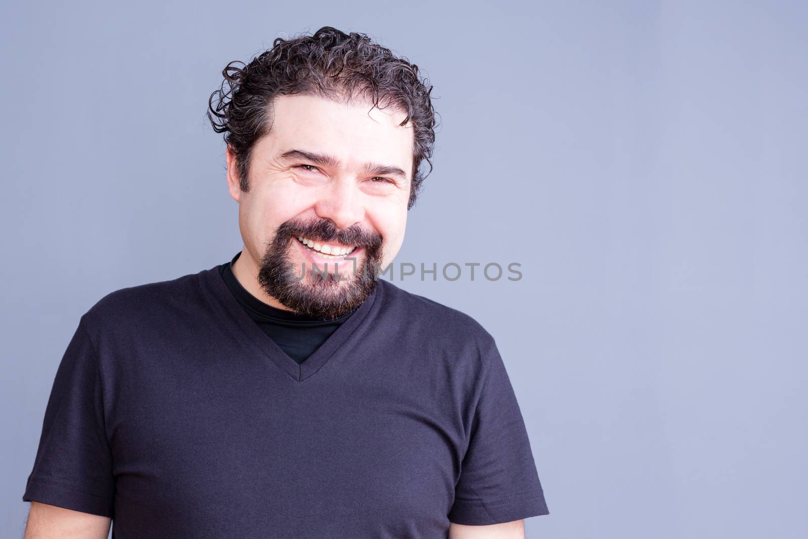 Waist Up Portrait of Mature Man with Beard and Dark Curly Hair Wearing Black T-Shirt and Laughing or Smiling at Camera in Studio with Gray Background and Copy Space