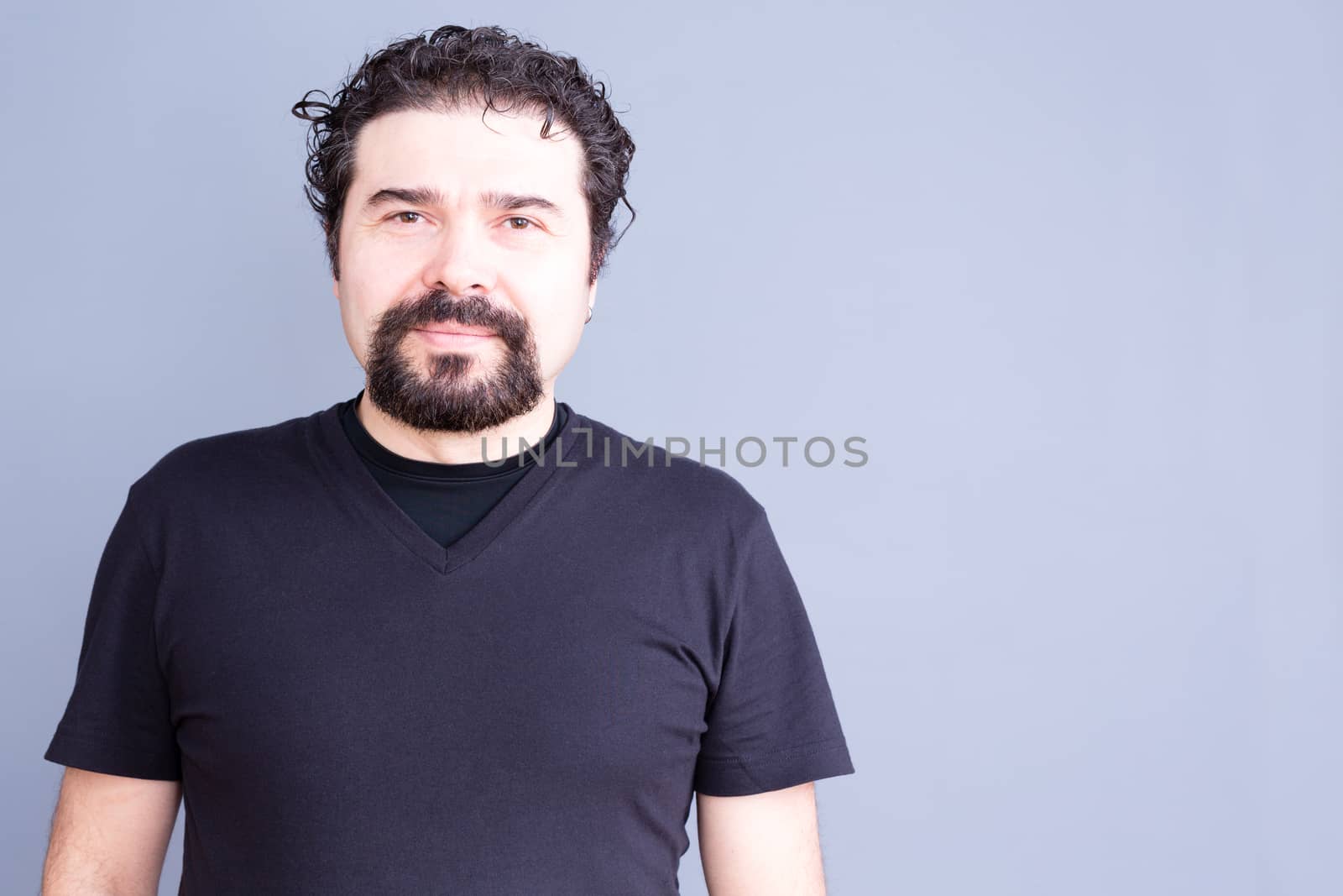 Waist Up Portrait of Mature Man with Beard and Dark Curly Hair Wearing Black T-Shirt and Staring at Camera with Blank Contented Expression, in Gray Studio with Copy Space