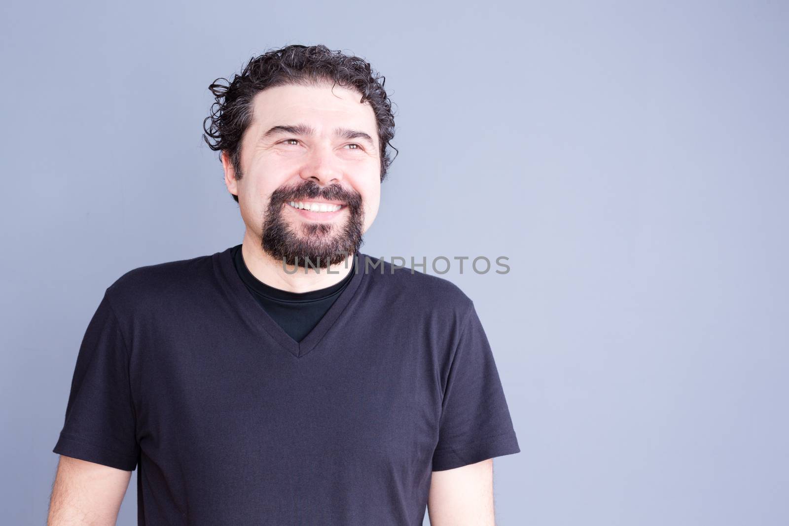 Waist Up Casual Portrait of Man with Beard and Curly Hair Wearing Dark T-Shirt Smiling and Laughing Joyfully in Studio with Gray Background and Copy Space