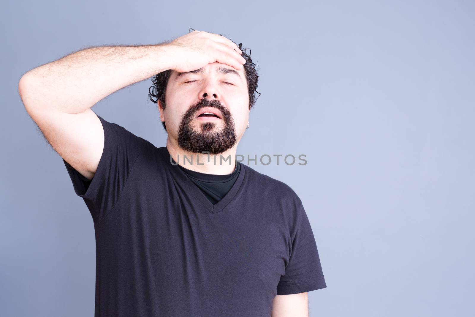 Waist Up of Frustrated Regretful Man with Beard Wearing Dark T-Shirt Holding Head and Hand on Forehead in Studio with Gray Background and Copy Space