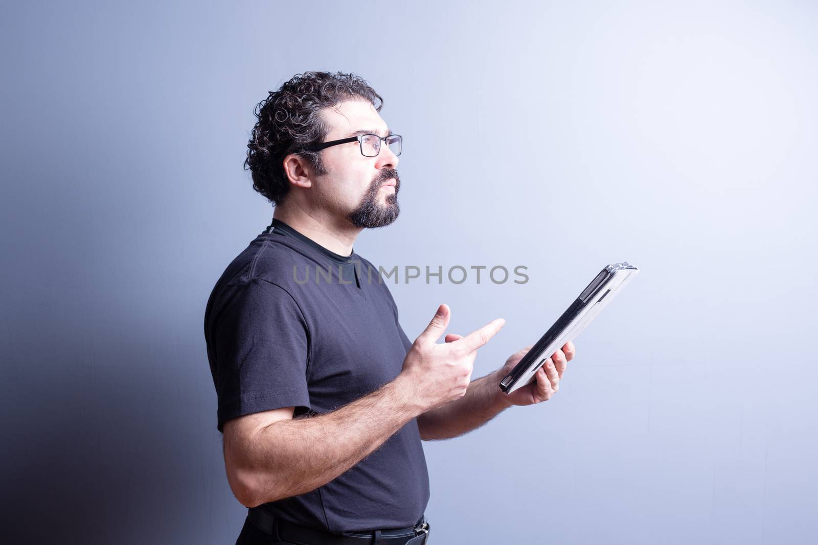 Profile of Man Wearing T-Shirt and Eyeglasses Deep in Thought While Holding Computer Tablet in Studio with Gray Background, Side Lighting and Copy Space