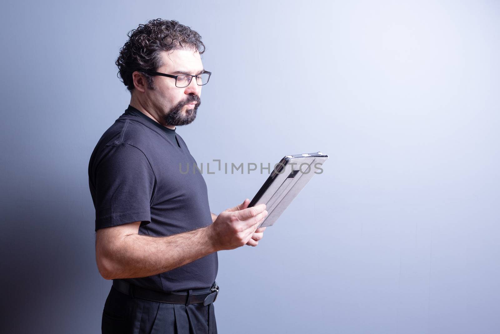 Profile of Man Wearing T-Shirt and Eyeglasses Looking Serious and Reading from Computer Tablet in Studio with Gray Background, Side Lighting and Copy Space