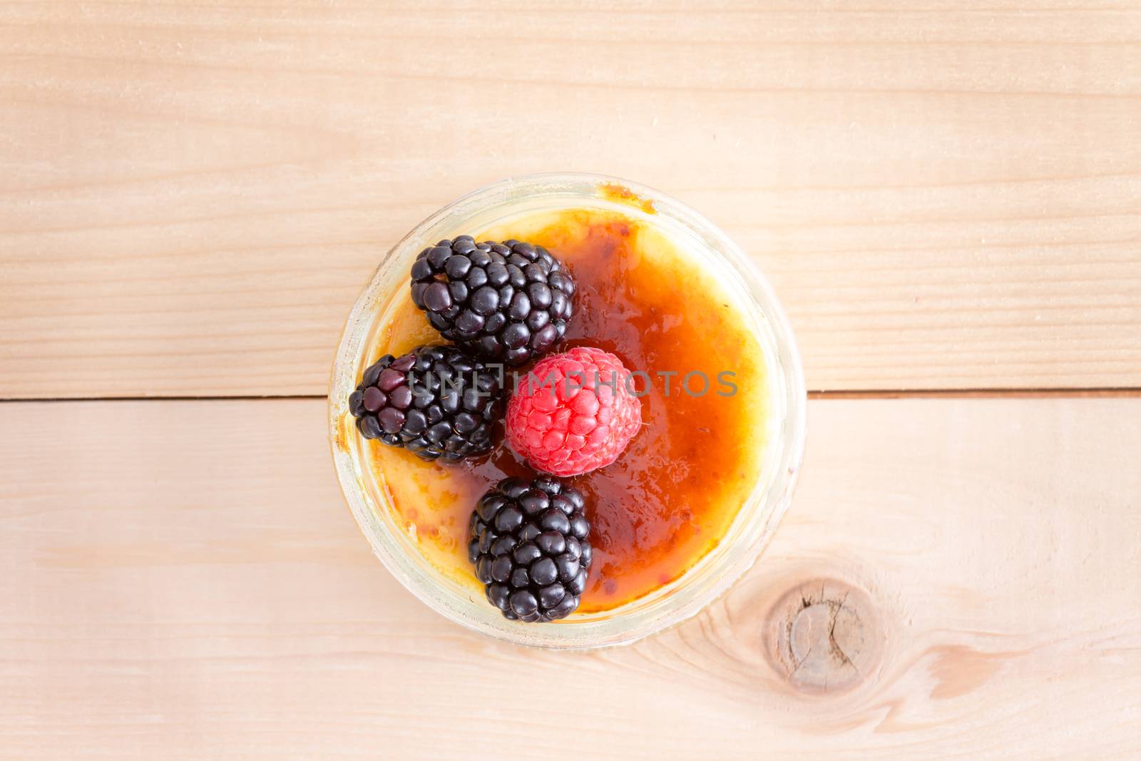 High Angle Still Life View of Single Serving Indulgent Creme Brule Custard Dessert Topped with Raspberry and Blackberry Fruit Garnish Served on Wooden Table Surface