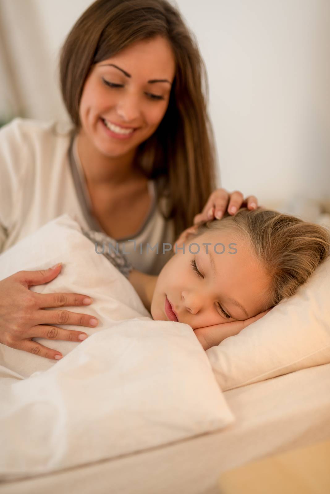 Happy attentive mother is lying in bed and hugging her sleeping daughter.