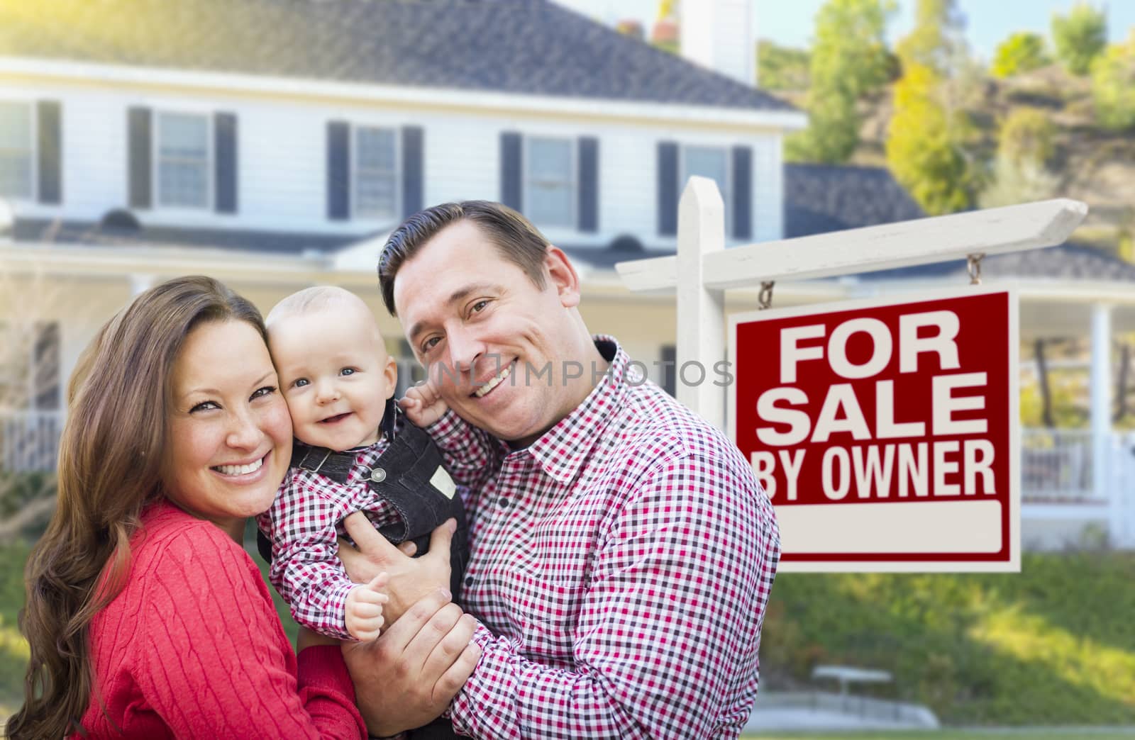 Happy Young Family In Front of For Sale By Owner Real Estate Sign and House.