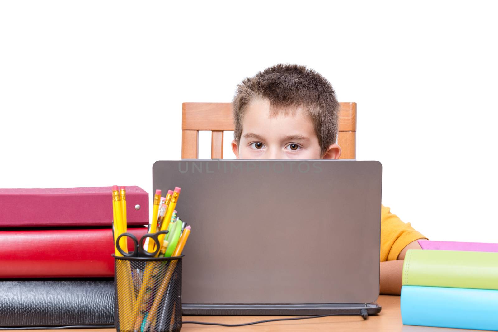 Young Boy Peeking Over Top of Laptop Computer Screen While Studying at Desk with Pencil Holder and Supplies and Surrounded by Colorful Books and Binders, in Room with White Background and Copy Space