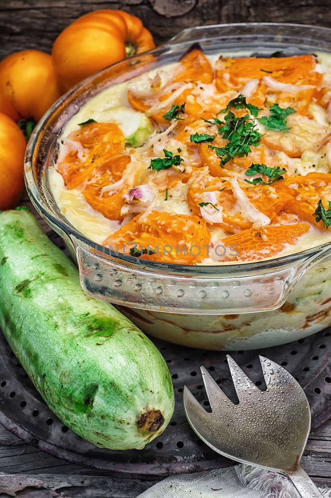 gratin of zucchini and yellow tomatoes on wooden background in glass bowl