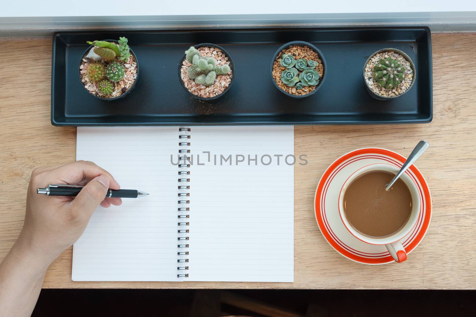 Top view of male hand writing on notebook and coffee cup on wood bar beside window in cafe in morning time