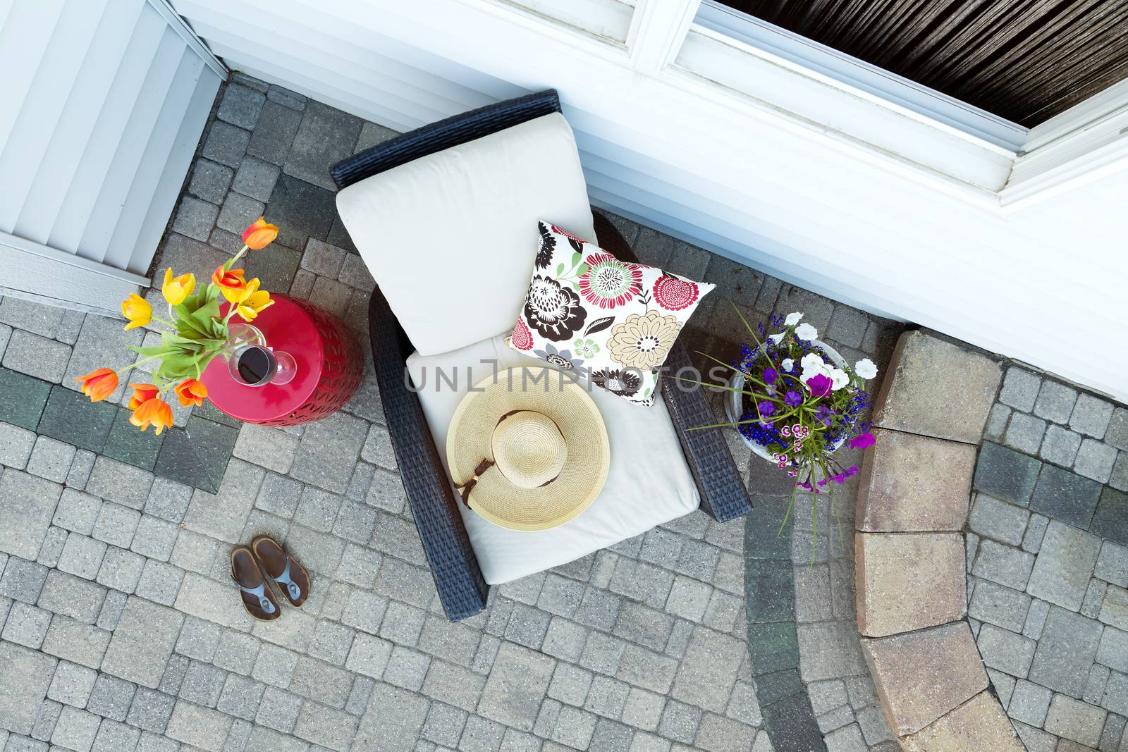 High Angle View of Sun Hat Resting on Comfortable Patio Chair with Floral Pattern Cushion and Framed by Colorful Flowers - Small Side Table with Glass of Red Wine and Shoes on Luxury Stone Patio