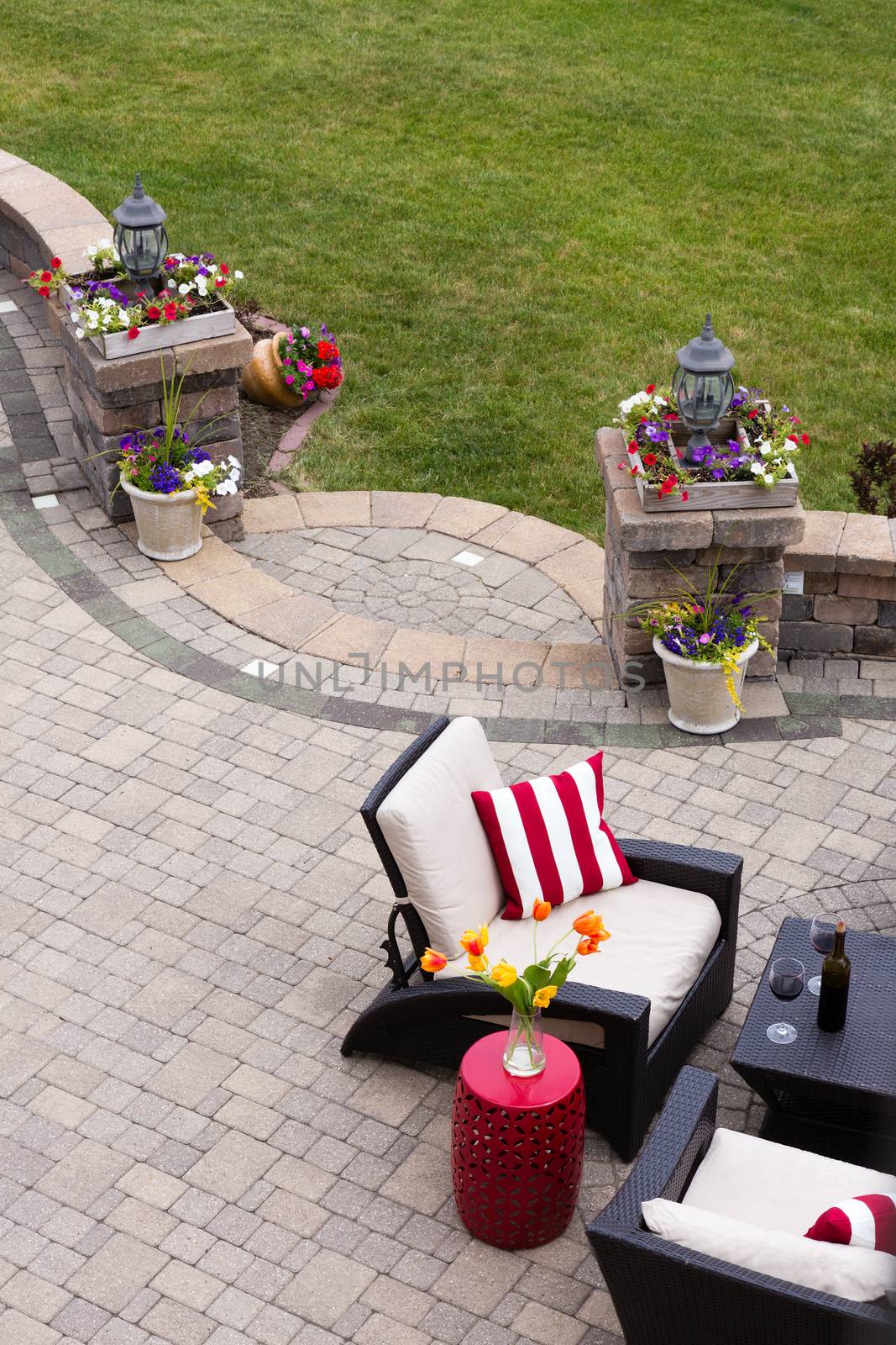 High Angle View of Luxury Patio with Outdoor Living Room Furniture and Stone Pillars Decorated with Colorful Flowers - Red Wine Served near Comfortable Chairs with Striped Cushions on Stone Patio