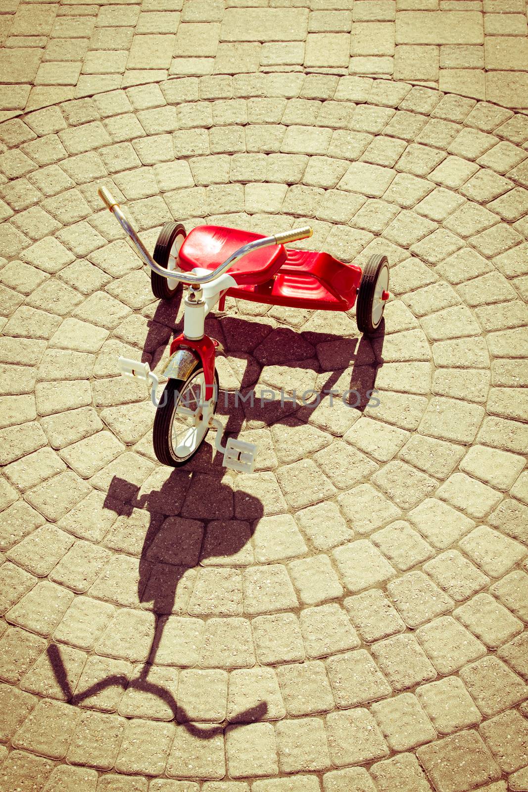 High Angle View of Red Vintage Tricycle in Bright Sunshine Casting Strong Shadow on Patio Stones Arranged in Circular Pattern Outdoors on Summer Day