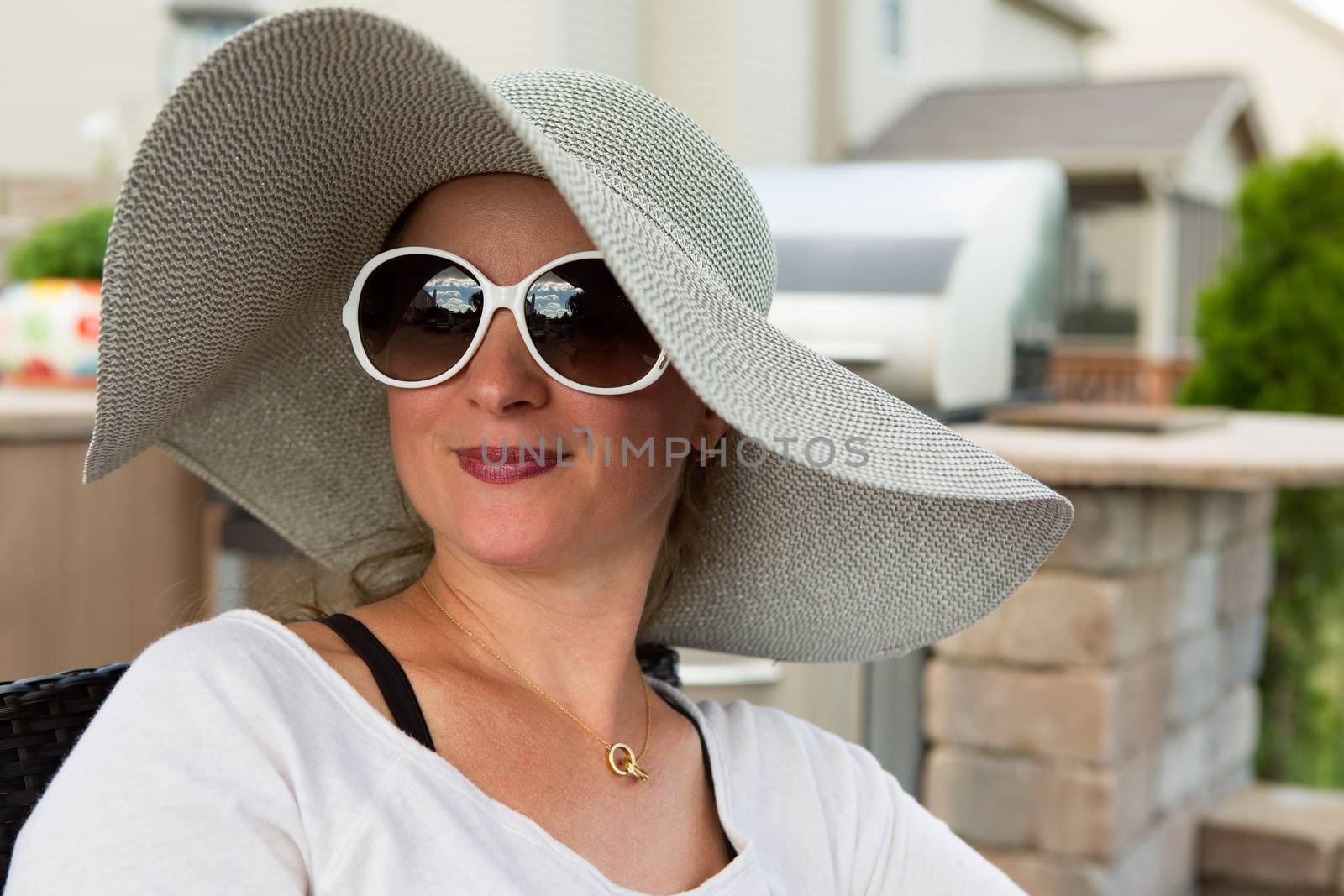 Head and Shoulders Close Up of Joyful Woman Wearing Large Brimmed Sun Hat and Sunglasses Smiling and Looking to the Side Outdoors on Backyard Patio on Sunny Day