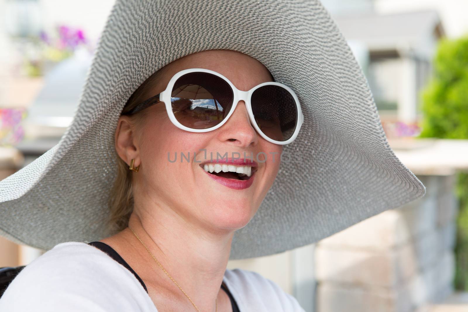 Head and Shoulders Close Up of Joyful Woman Wearing Large Brimmed Sun Hat and Sunglasses Laughing Outdoors on Backyard Patio on Sunny Day