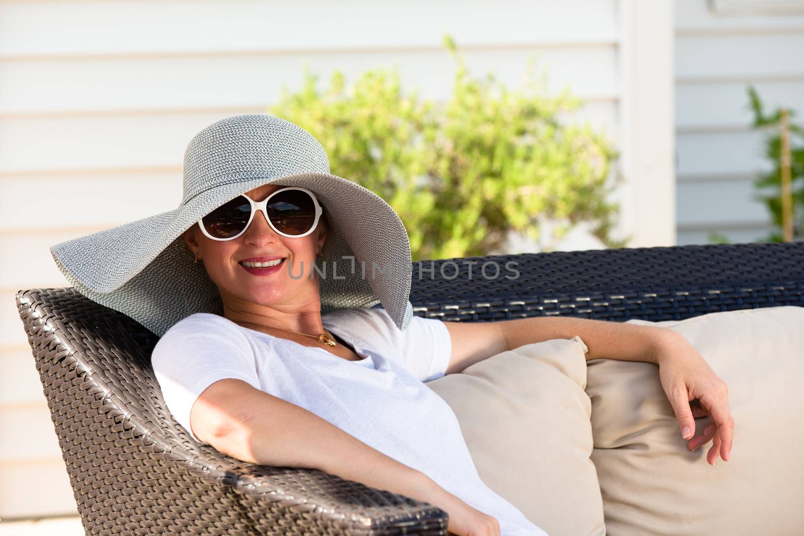 Candid Waist Up Portrait of Woman Wearing Large Brimmed Sun Hat and Sunglasses Relaxing Outdoors on Patio Love Seat with Comfortable Cushions in Back Yard on Sunny Summer Day