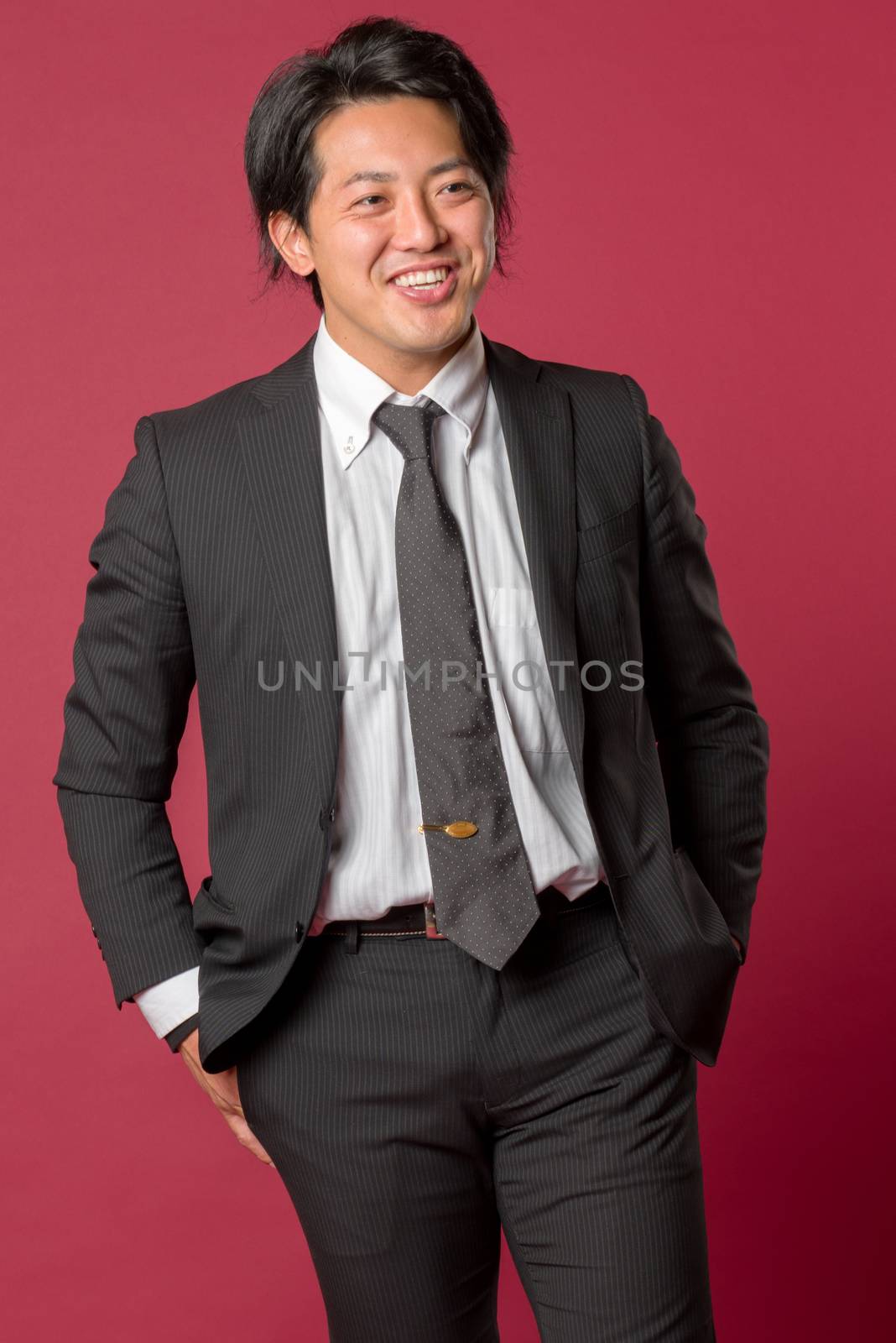 A half body portrait of a young Japanese man in a business suit laughing on a red background.
