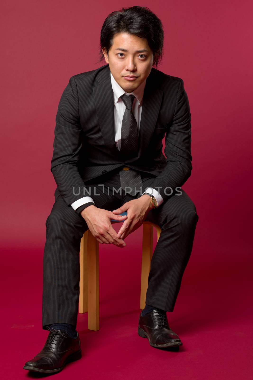 A portrait of a young Japanese man in a business suit sitting on a stool in a cool pose.
