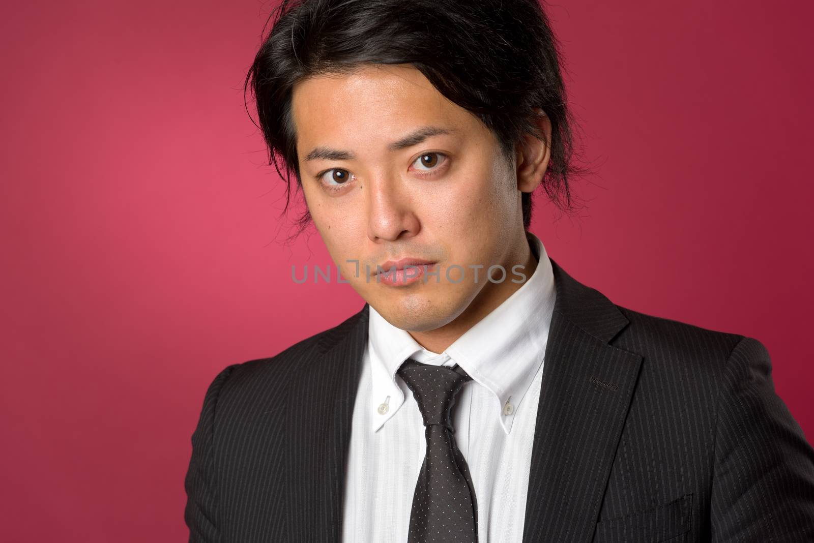 A headshot style portrait of a young Japanese man wearing a business suit with a serious expression on a red background.