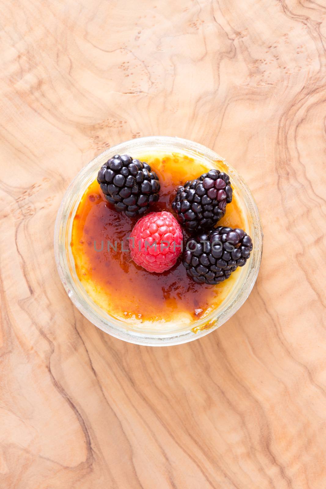 Above view of single little bowl full with custard dessert and toppings of black and red raspberries over wooden surface