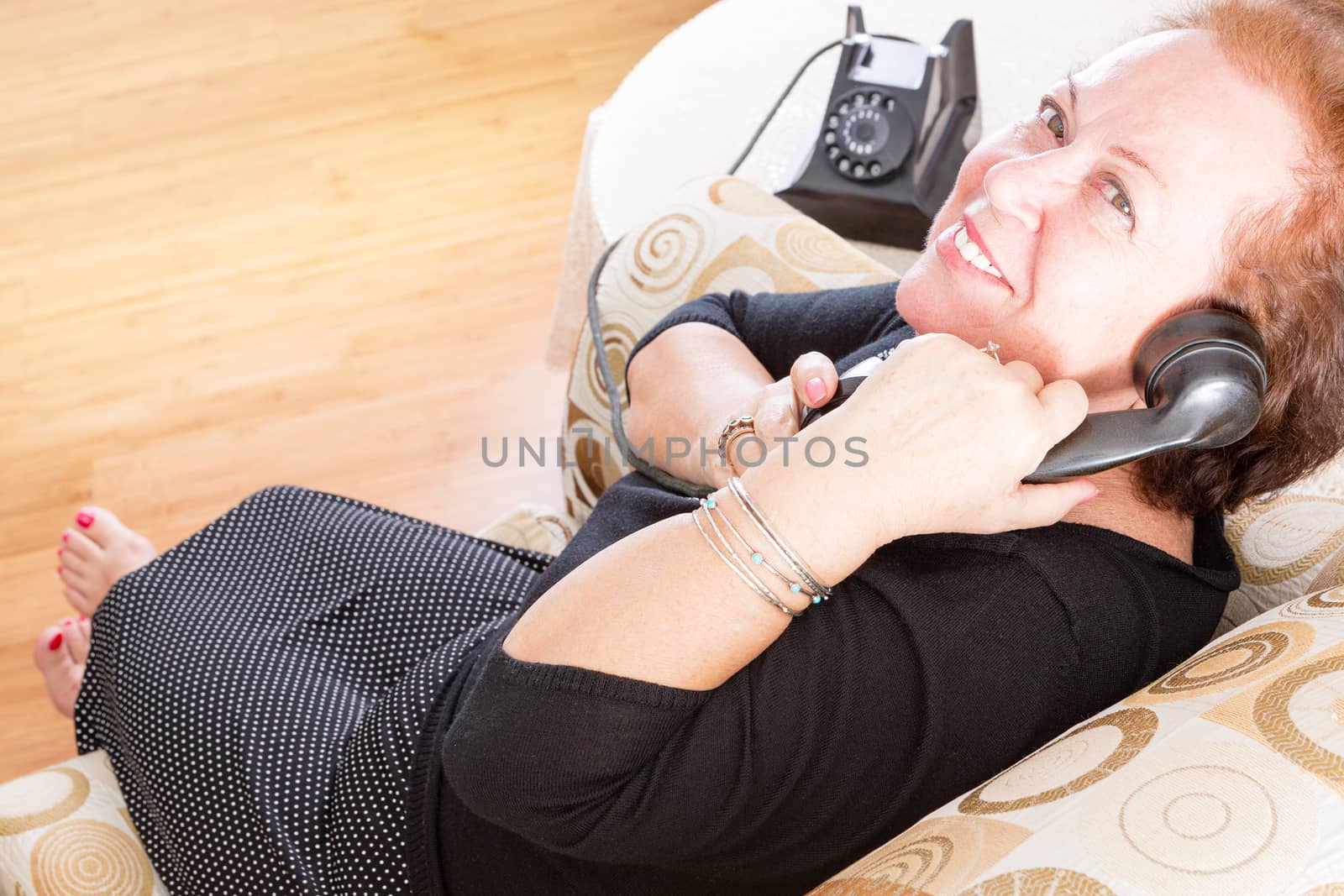Grandmother relaxing at home in a comfy armchair in a stylish black dress and bare feet talking on a retro rotary phone looking up with a smile to confirm good news, view from above
